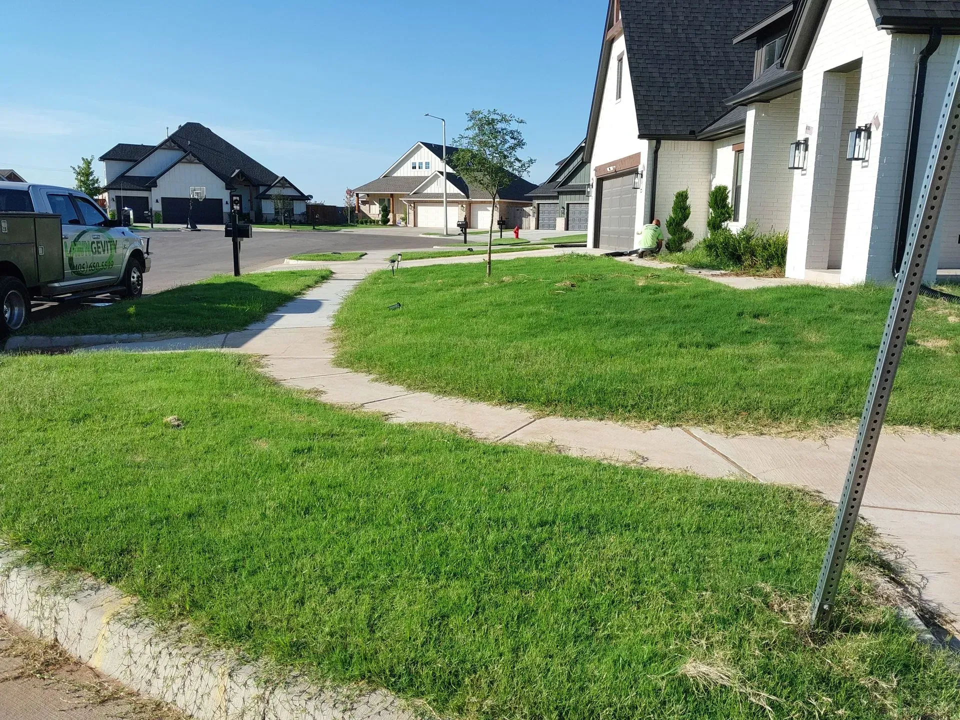 Green lawn next to sidewalk in front of houses with dark roofs on a sunny day.