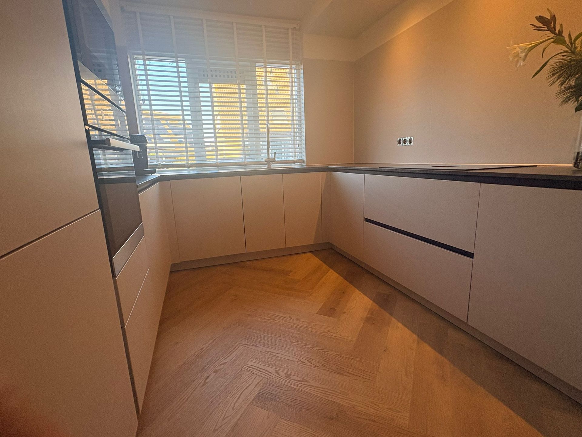 Beige keuken met houten vloer, kasten, donker aanrechtblad en raam met jaloezieën.