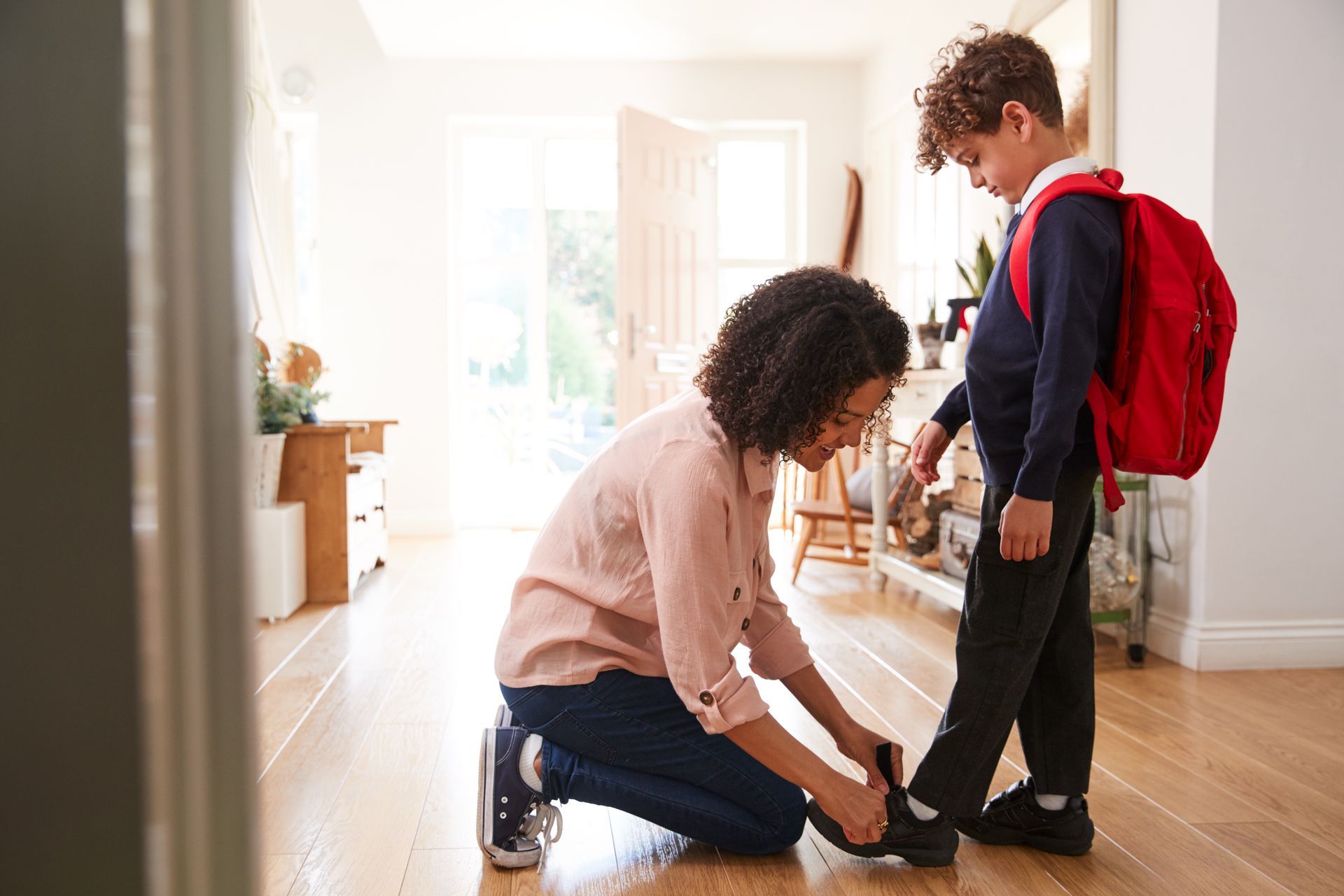 Adult kneeling to tie a child’s shoe in a bright hallway, with a red backpack nearby.