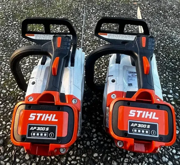 Two Stihl battery-powered chainsaws with orange and black accents, sitting on a rough surface.