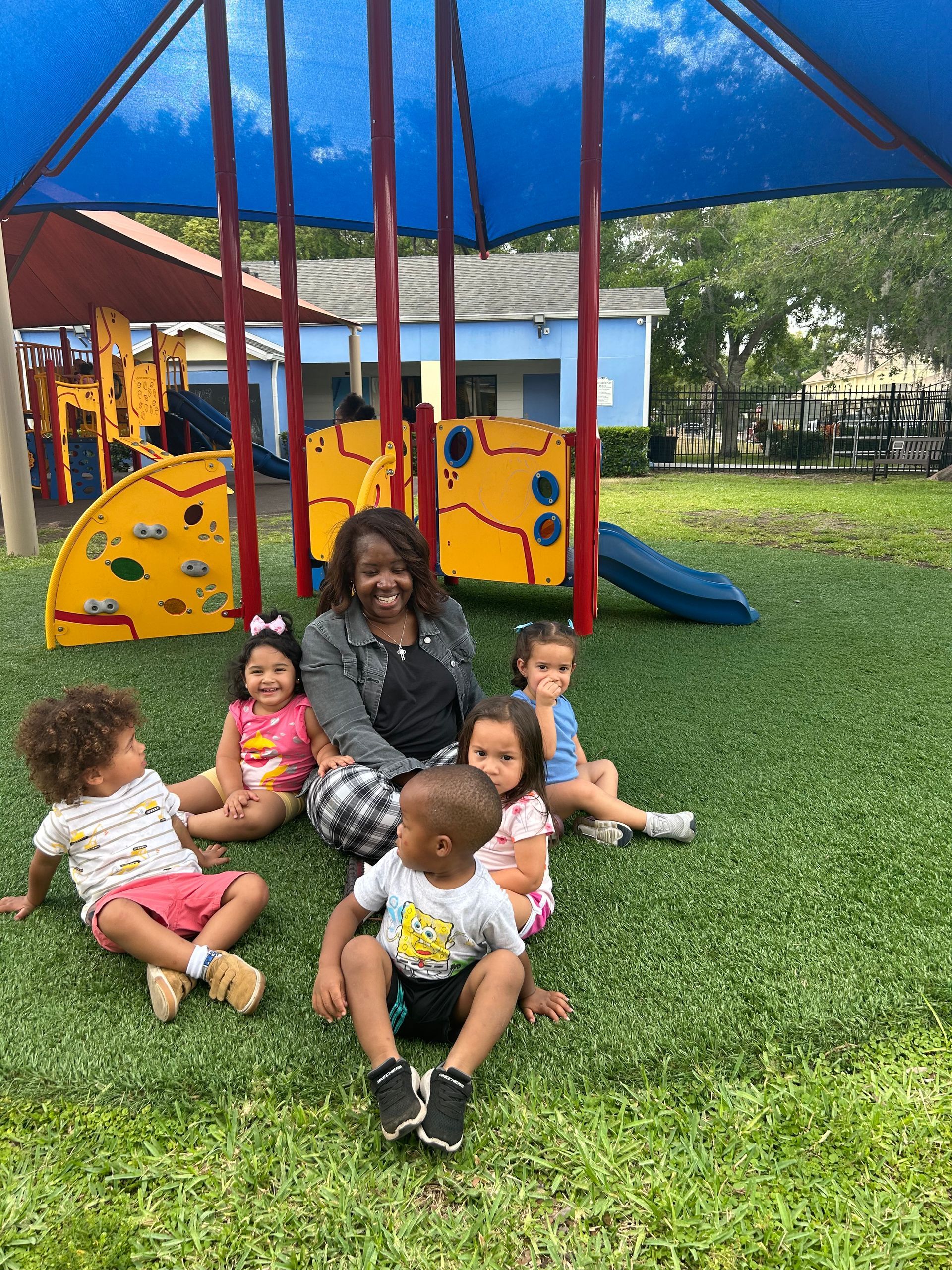 A woman is sitting on the grass with a group of children in front of a playground.