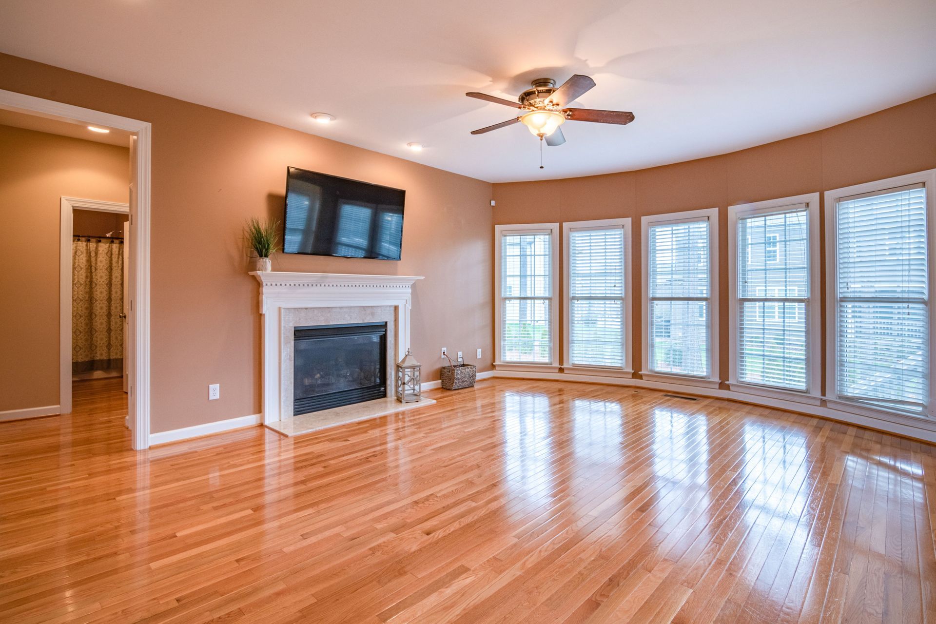 Bright living room with hardwood floors, fireplace, wall-mounted TV, and large windows with blinds