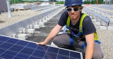 A man wearing a hard hat and sunglasses is kneeling next to a solar panel.