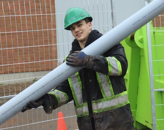 A man wearing a green hard hat is carrying a large pipe