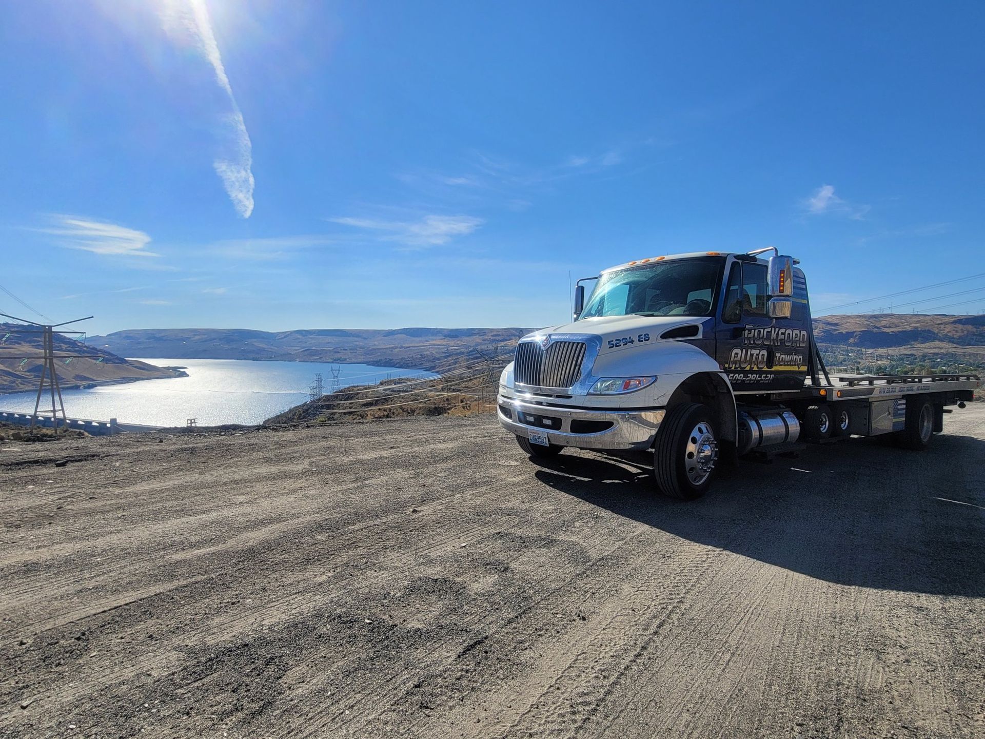 A tow truck is parked on a dirt road next to a lake. | Rockford Auto Inc