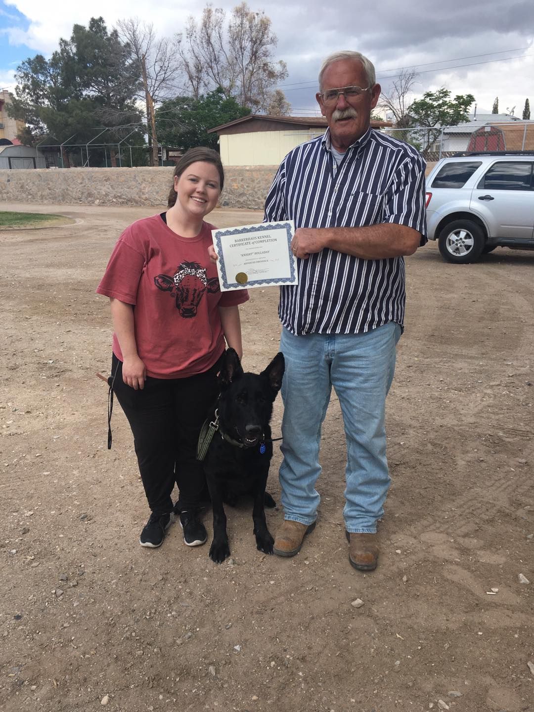 A woman and man with a black dog, holding a certificate. Outdoors on dirt.