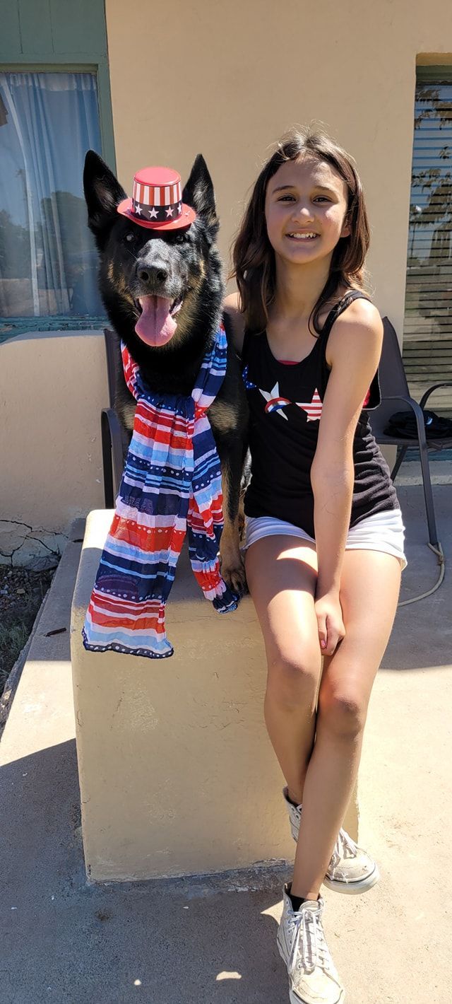 A young girl and a dog in patriotic outfits posing outdoors. Girl smiles next to the dog wearing a hat and pants.