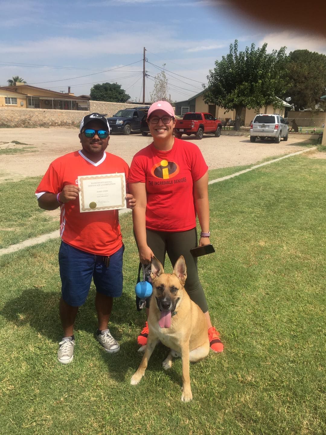 Two people and a dog posing for a photo outdoors. One person holds a certificate.