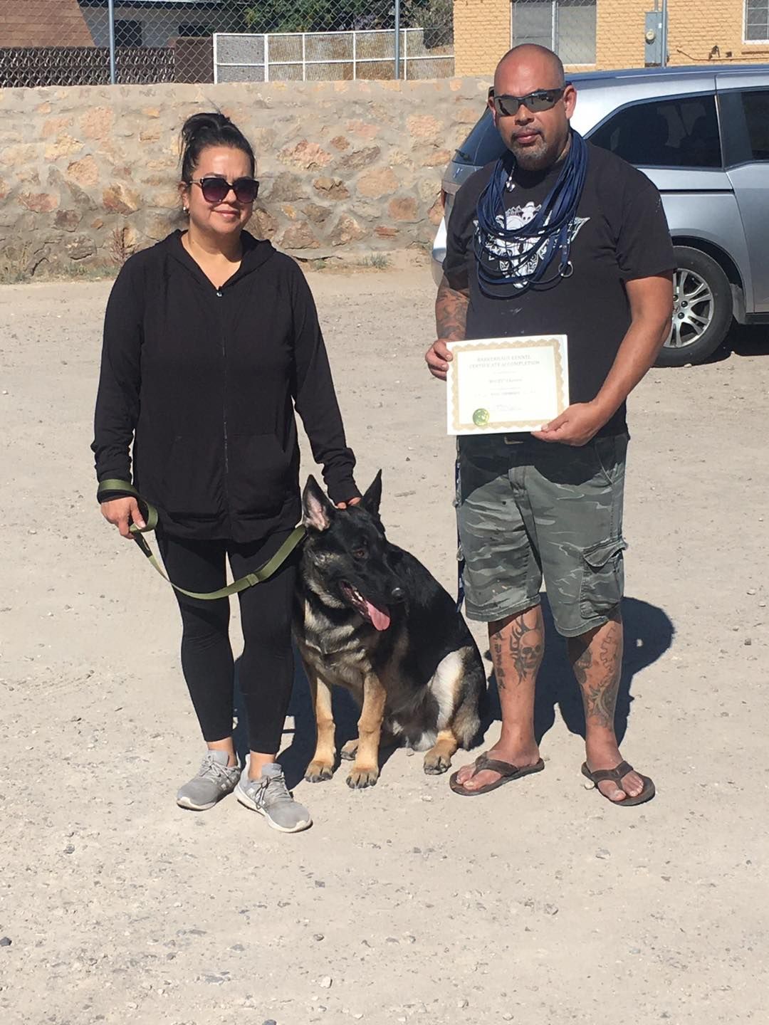 A woman, man, and German Shepherd with a certificate. Outdoors. Man holds the certificate.