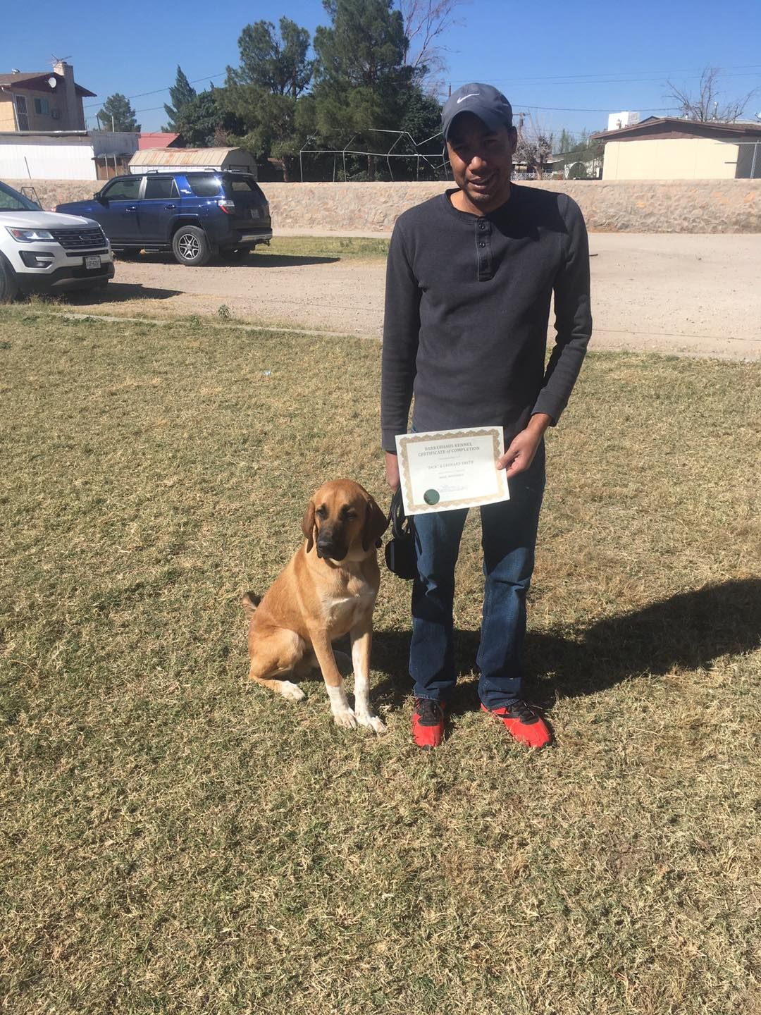 Man and dog seated on grass, man holding certificate. Blue jeans, red shoes, dog is brown.
