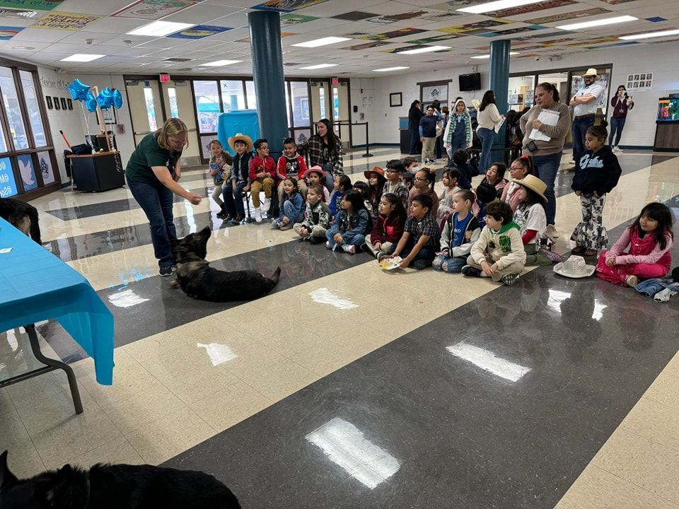 A woman with a dog demonstrating to a group of seated children in a school hallway.