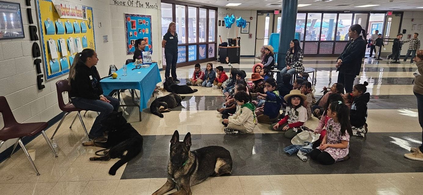 Children and dogs at an event in a school hallway. Adults and dogs are present. Children are seated on the floor.
