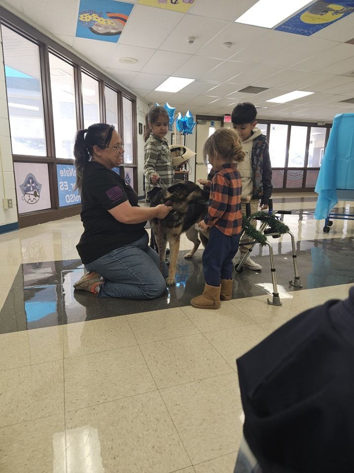 Woman kneeling petting a dog while children watch in a school hallway.
