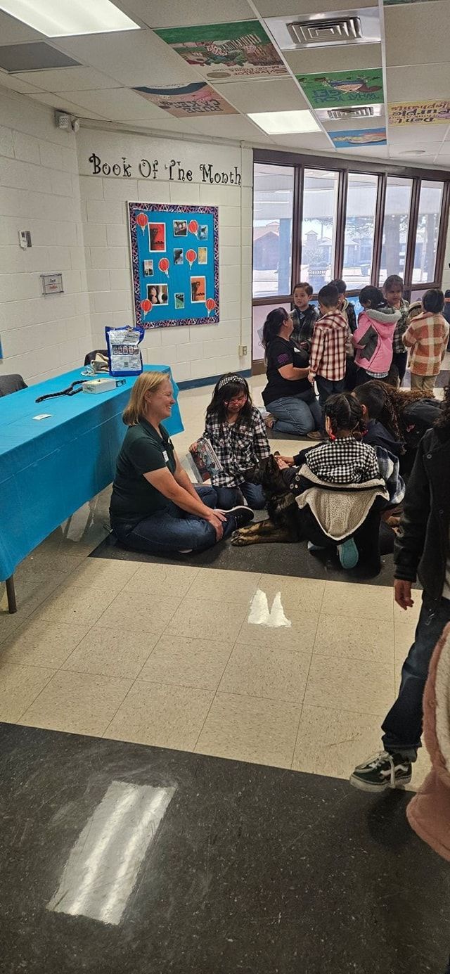 Teacher and students in a classroom, some seated on the floor. One person kneels on a mat, near two cups.