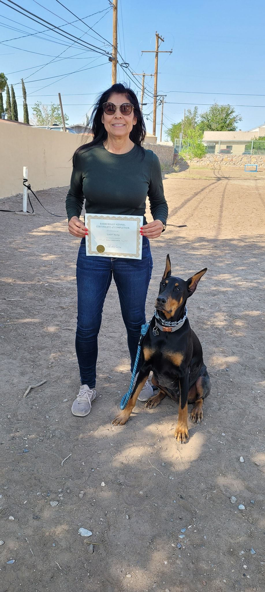 Woman with dog poses outside holding a certificate. The dog is black and tan with its ears cropped.