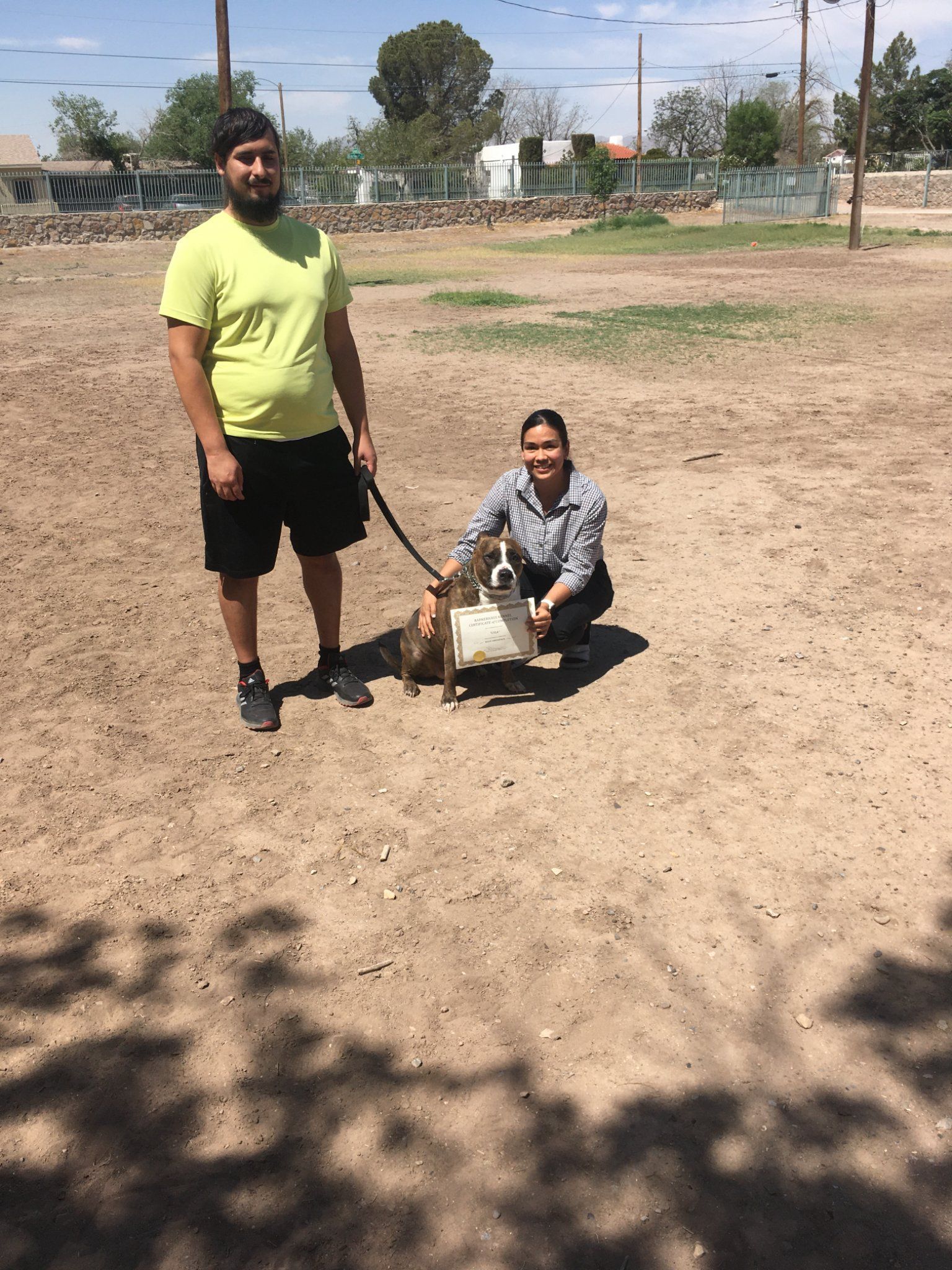 A man and woman with a dog in a dirt field on a sunny day.