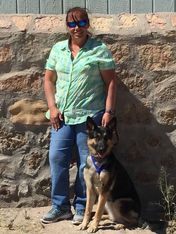 Woman in green shirt and jeans, standing with a German Shepherd dog in front of a stone wall.