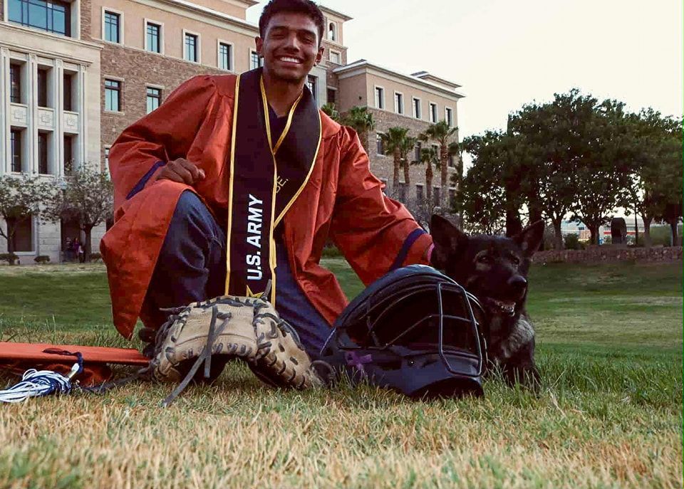Man in graduation gown kneels with gloves and dog on grass, U.S. Army sash, building in background.