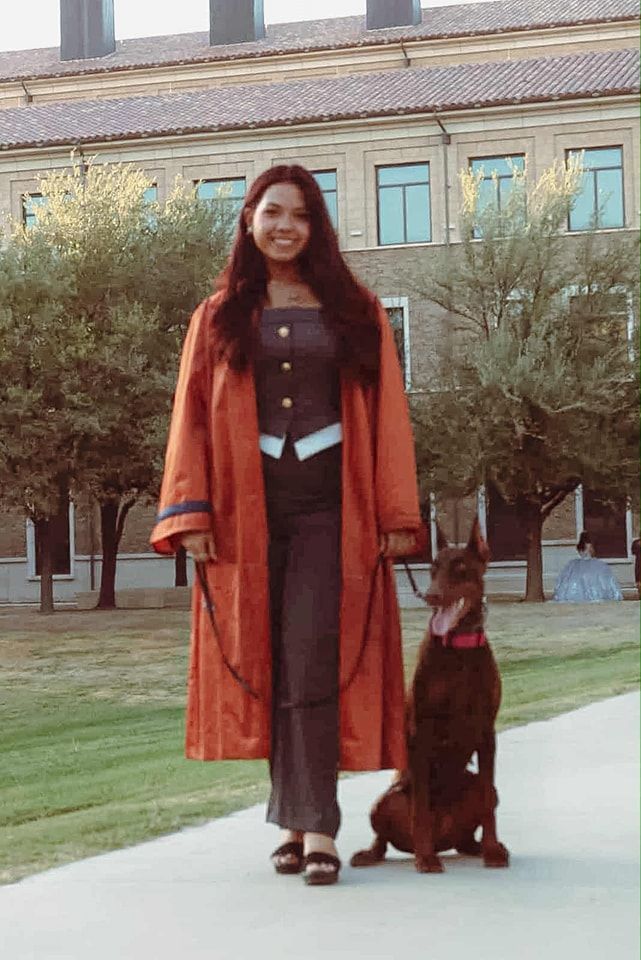 Woman in red coat and suit stands with brown Doberman in front of a brick building.
