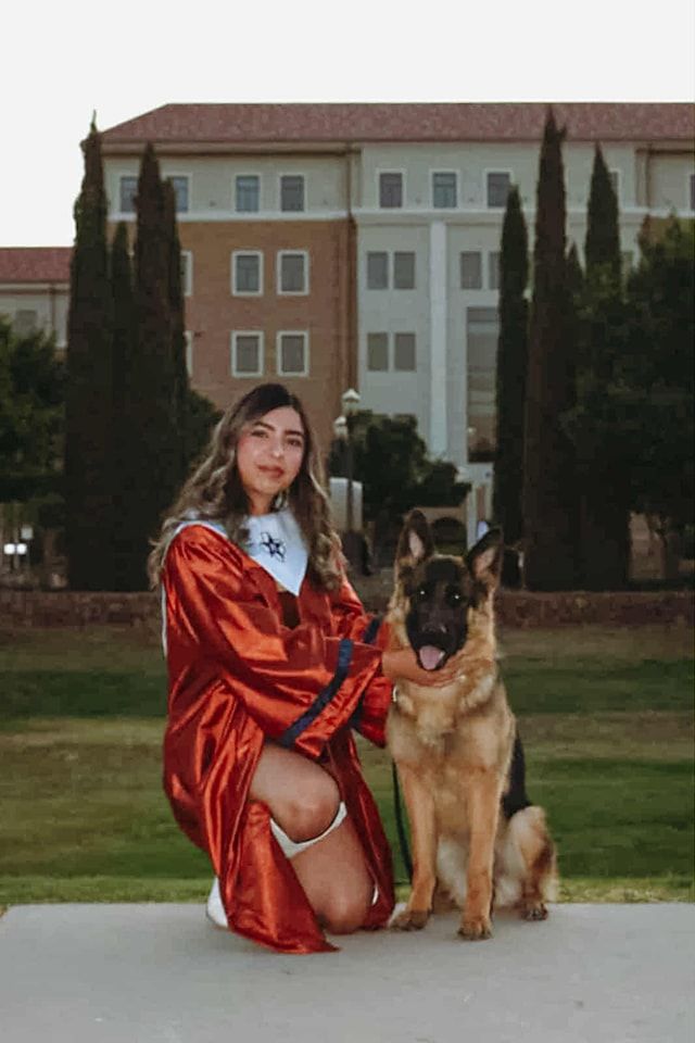 Woman in graduation gown kneels with German Shepherd in front of a building.
