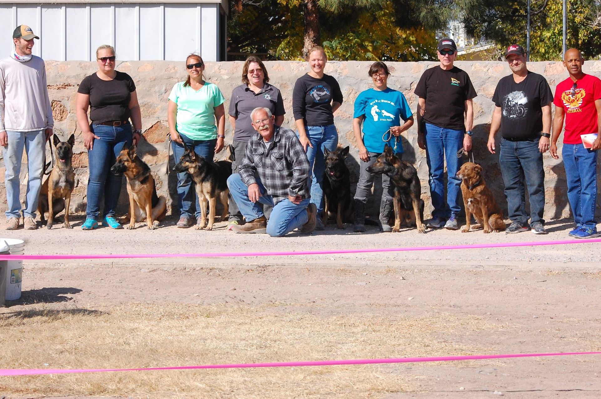 Group of people and dogs posing outdoors, on a sunny day. They stand behind a pink ribbon.