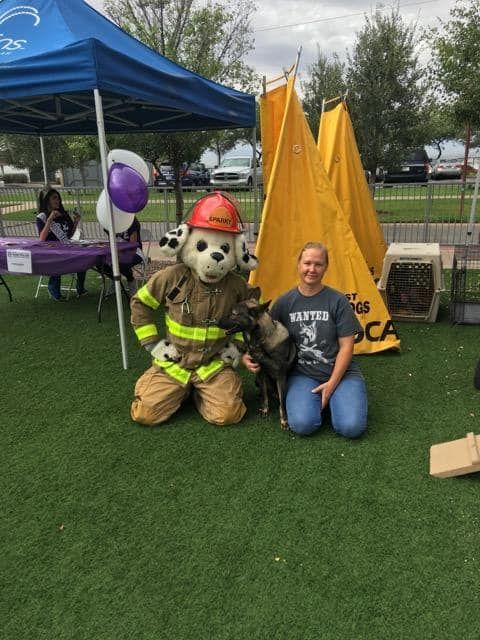 A firefighter mascot kneels with a woman and dog at an event with tents and tables.
