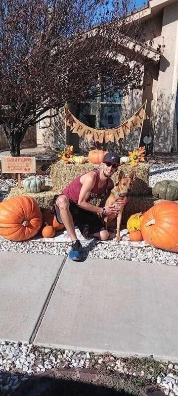 Man with dog poses in front of pumpkins and hay bales, celebrating fall outside a house.