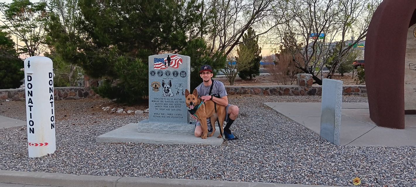Person with a dog kneeling by a veterans memorial. Setting is outdoors in front of the monument.