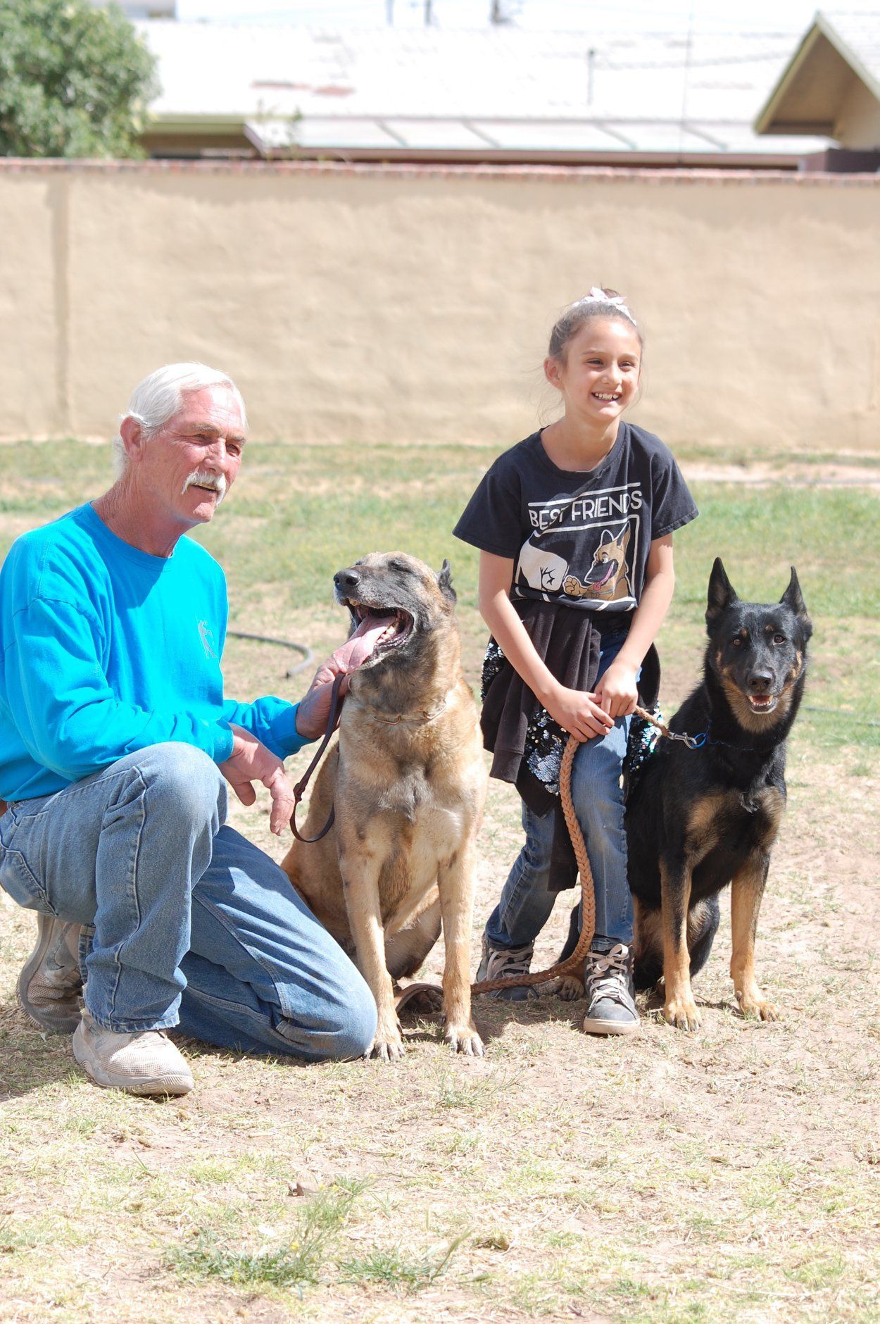 Man and girl with two dogs outdoors; man kneels, girl stands, dogs sit.