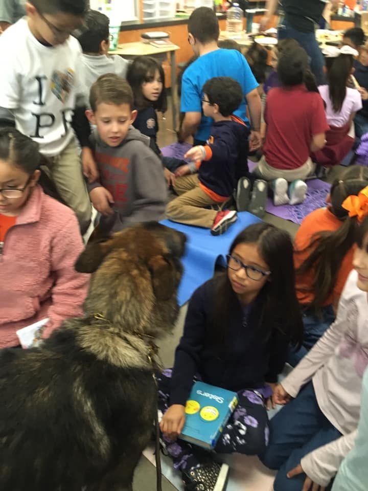Students and a German Shepherd in a classroom. Children read while the dog sits with them.