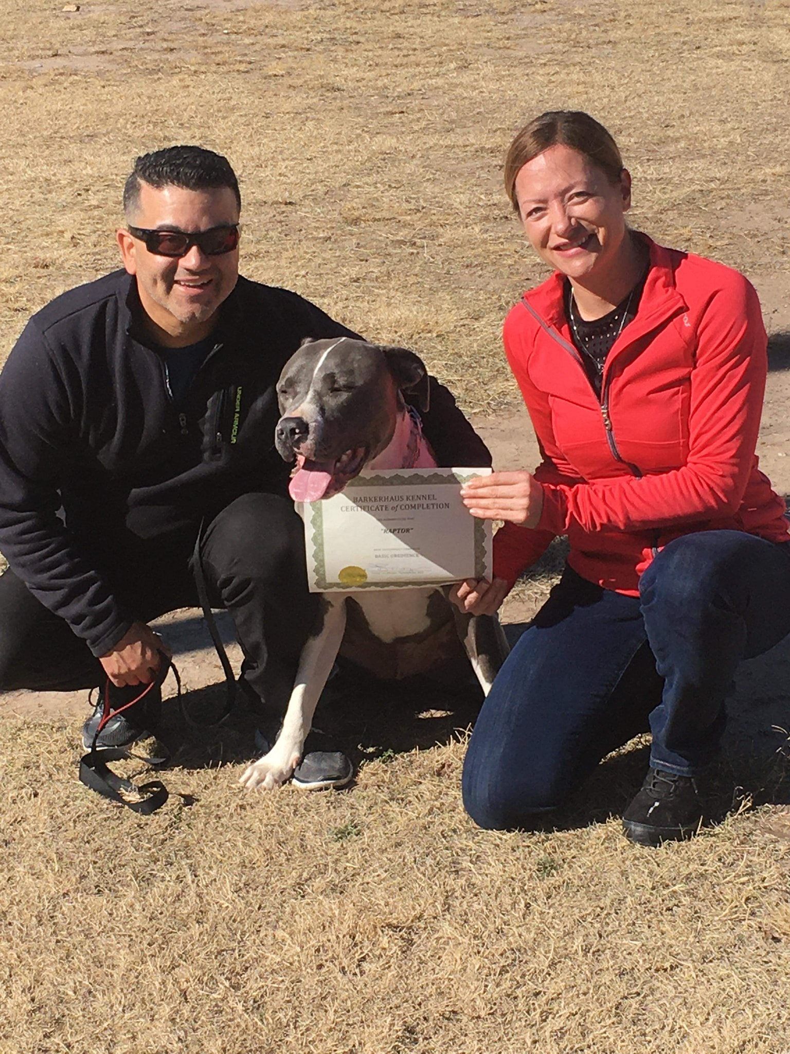 Man and woman kneel with a pit bull dog, holding a certificate outdoors.