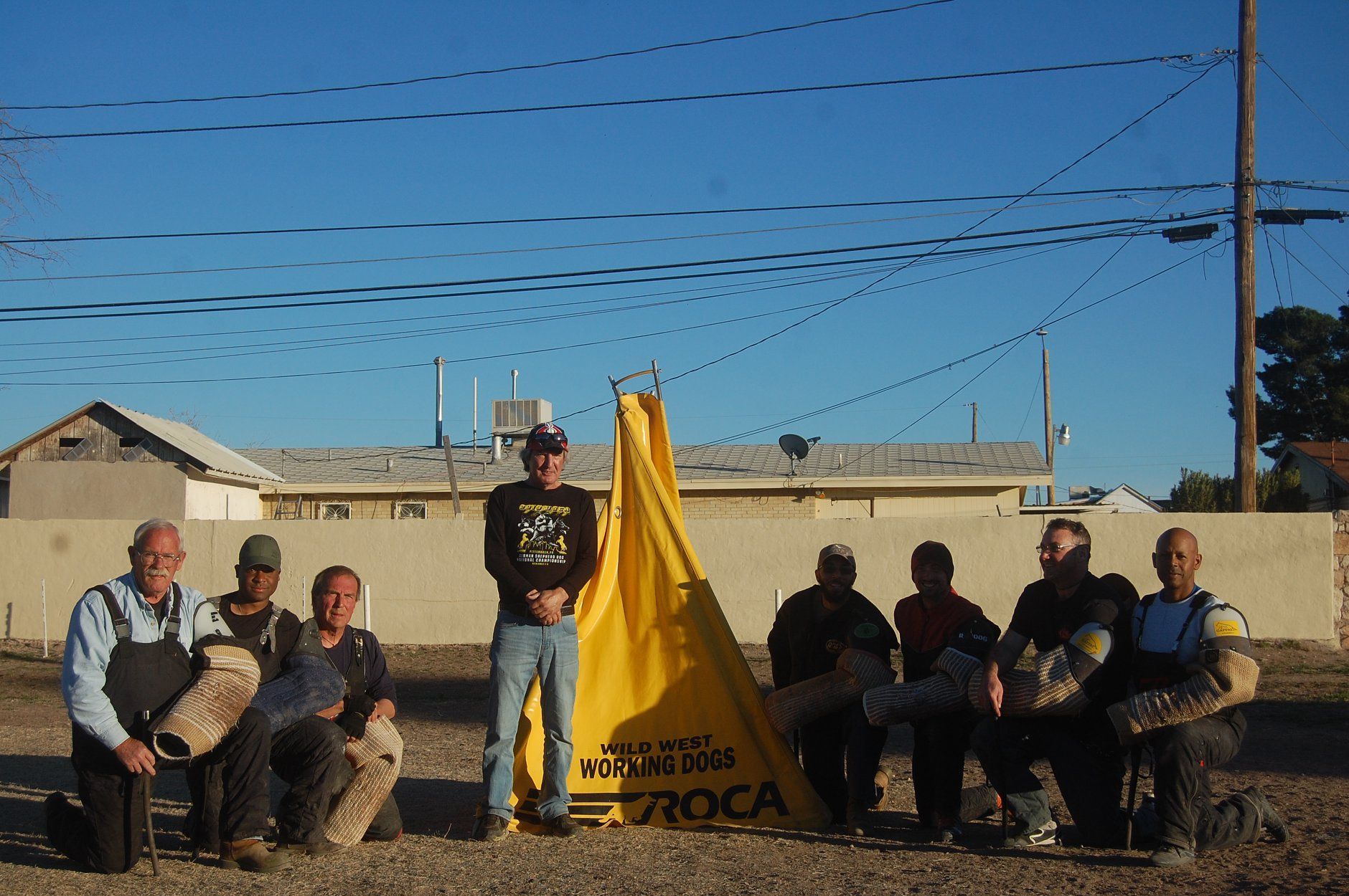 Group of men posing near a yellow tent; outdoor setting; blue sky and buildings in background.
