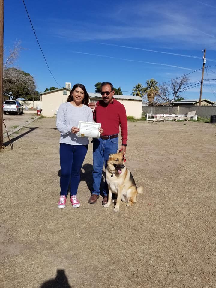 Woman and man with a dog, holding a certificate outside on a sunny day.