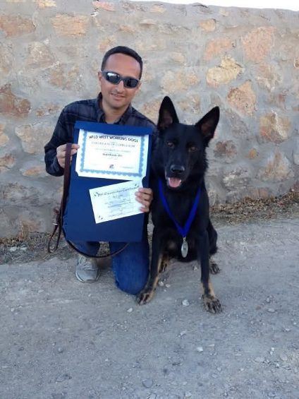Man and black dog kneeling, holding awards. Dog wears a medal. Outdoor setting, stone wall.