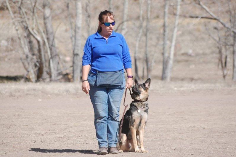 Woman in blue top, sunglasses, jeans with a German Shepherd dog on a leash in an outdoor setting.