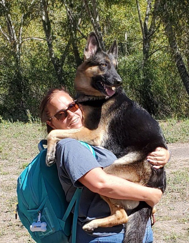 Woman with sunglasses hugs a large black and tan German Shepherd outdoors, blue backpack visible.