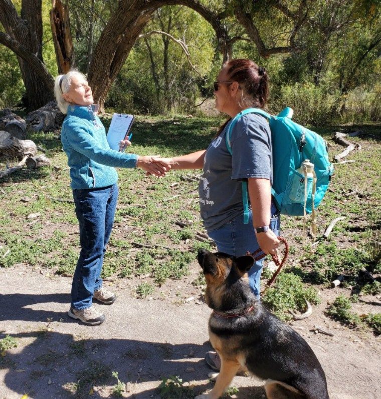 Two women shaking hands in a park; one holds a clipboard, the other has a backpack and dog.