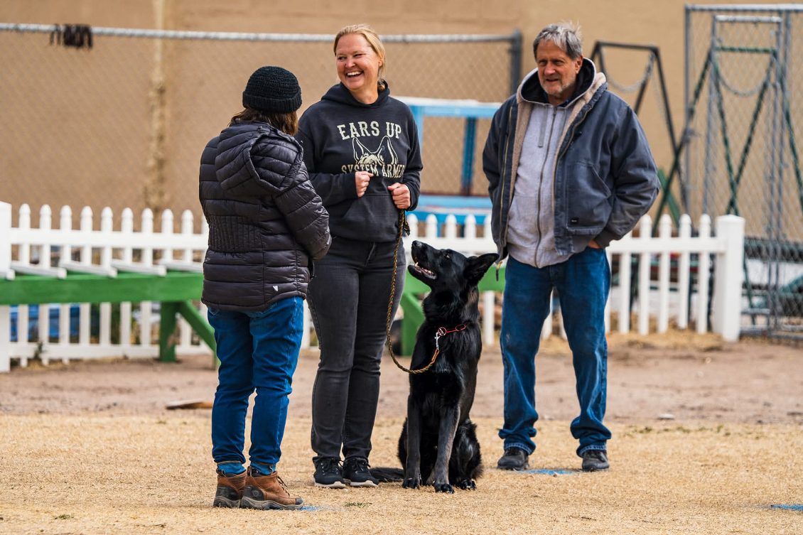Three people with a black dog outside. Woman in black jacket and jeans watches dog. Woman in black hoodie smiles. Man in blue jeans smiles.