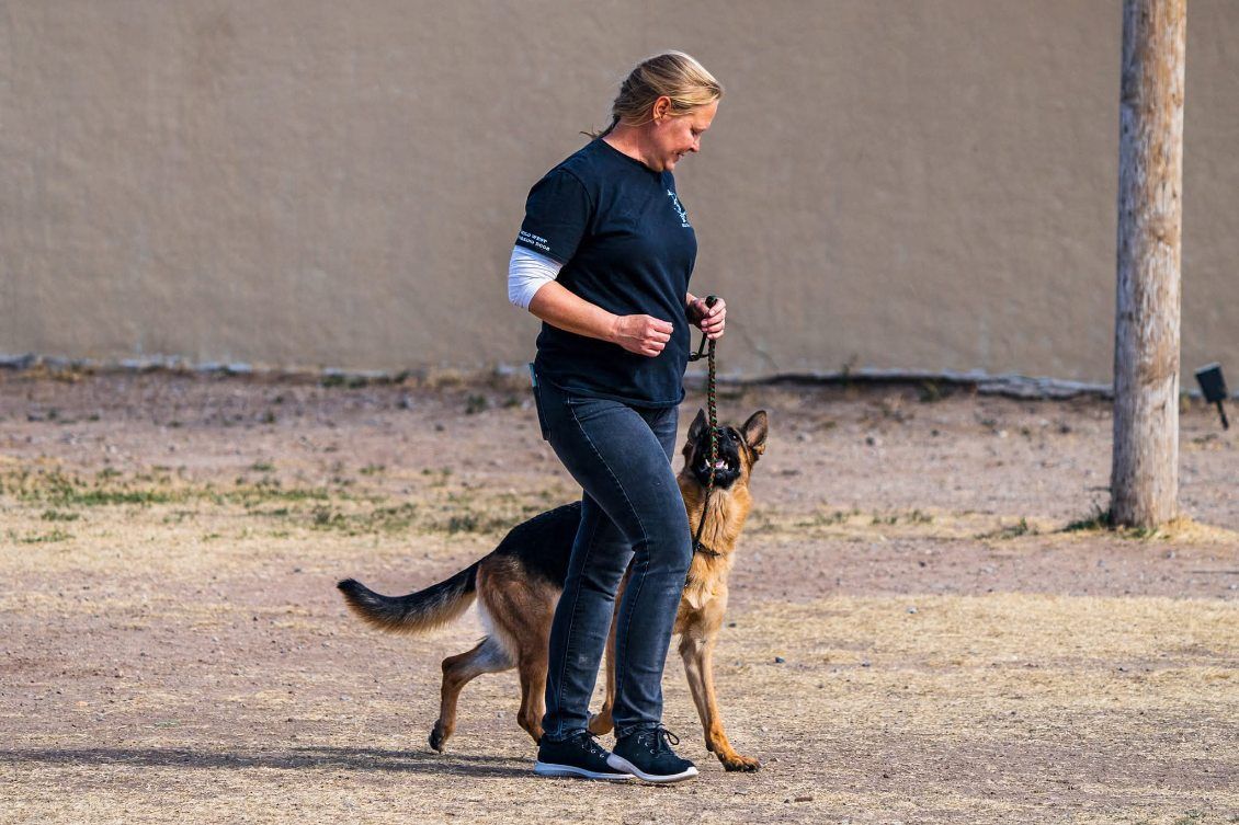 Woman training a German Shepherd outdoors; both facing forward. The dog is leashed and focused.