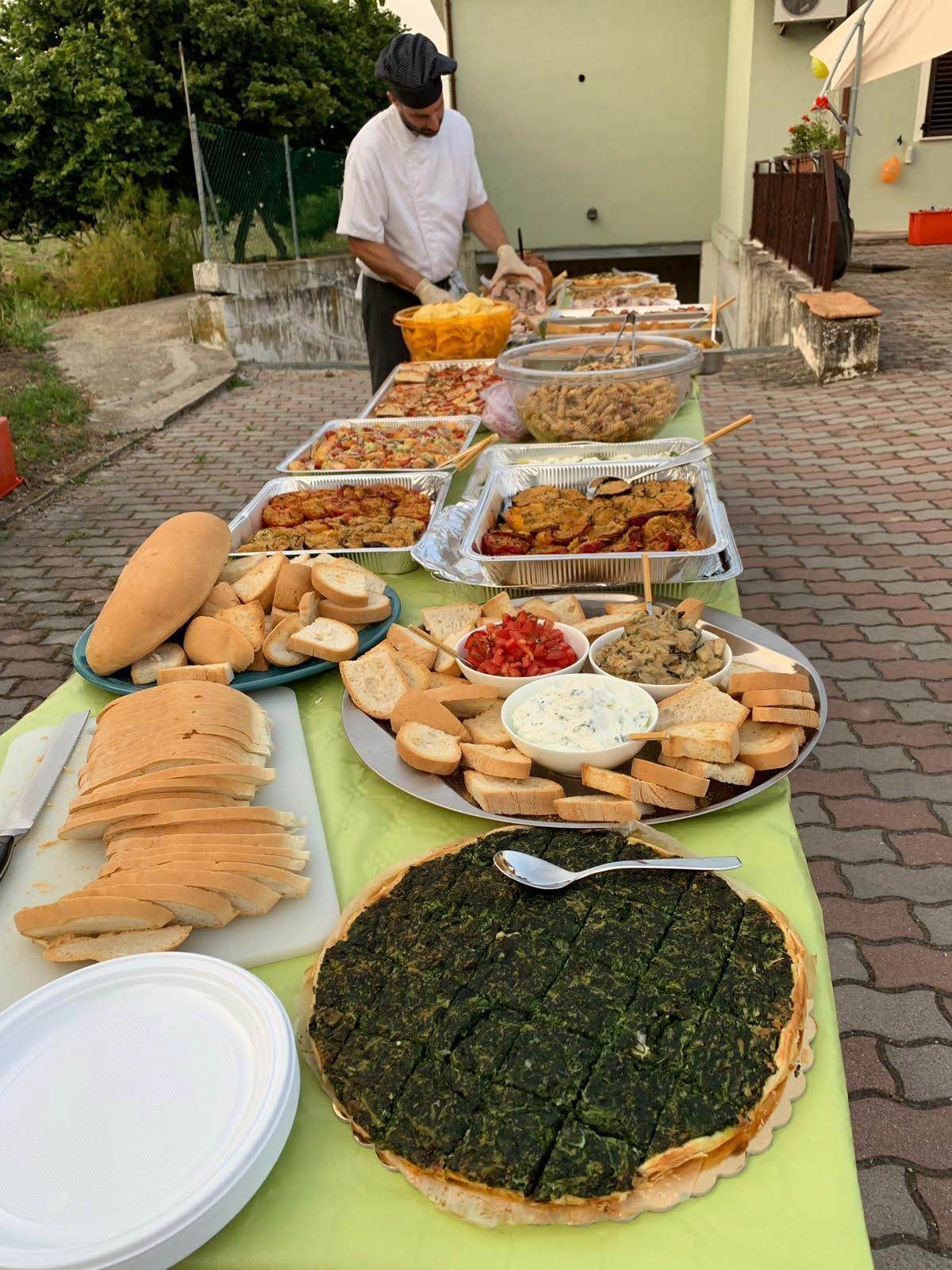 Tavolo da buffet all'aperto con pane assortito, salse e piatti grigliati; una persona è in piedi dietro di esso in un cortile.