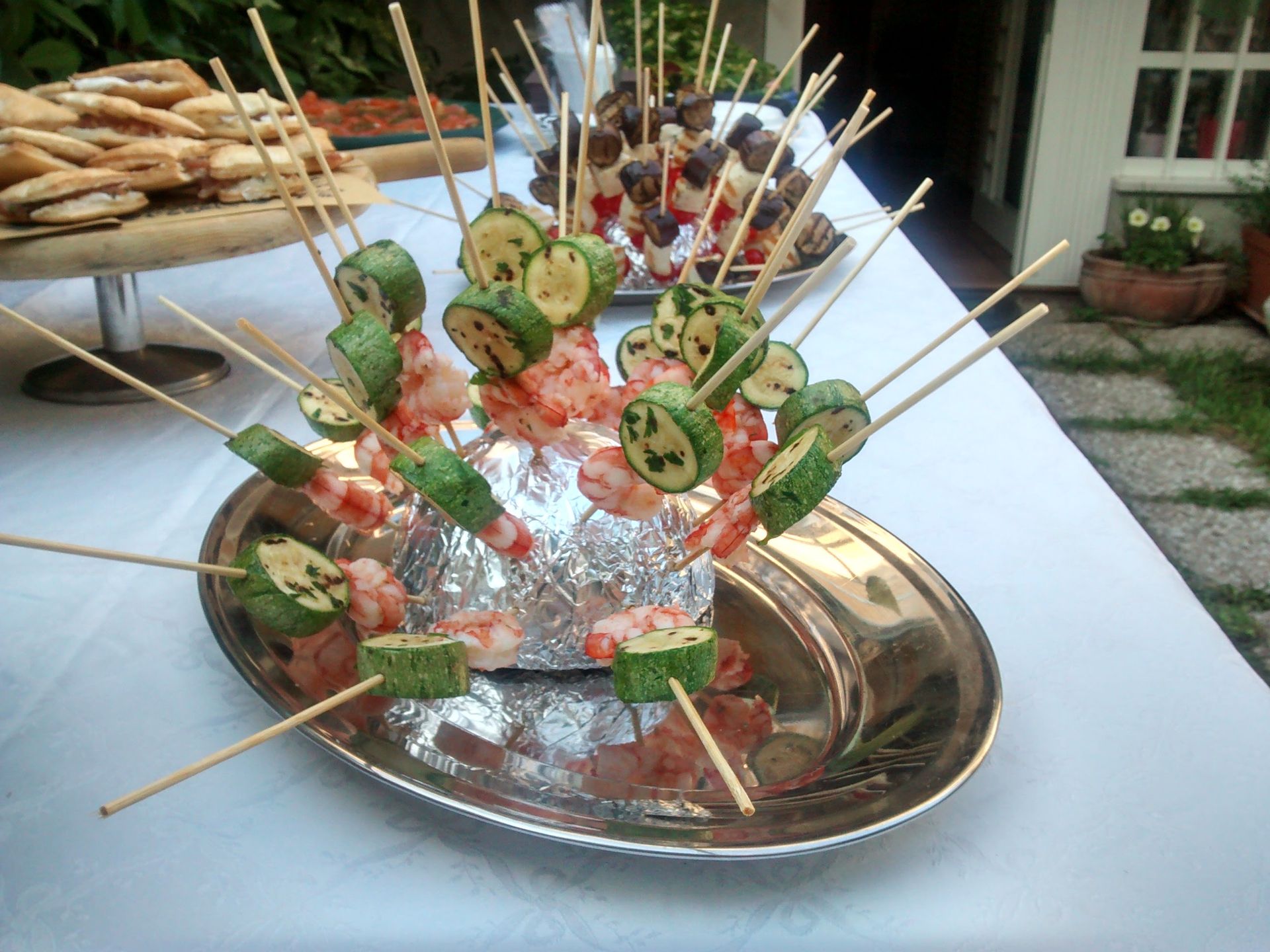 Antipasti infilzati su spiedini e disposti in un piatto circolare decorativo con erbe aromatiche fresche e guarnizioni.