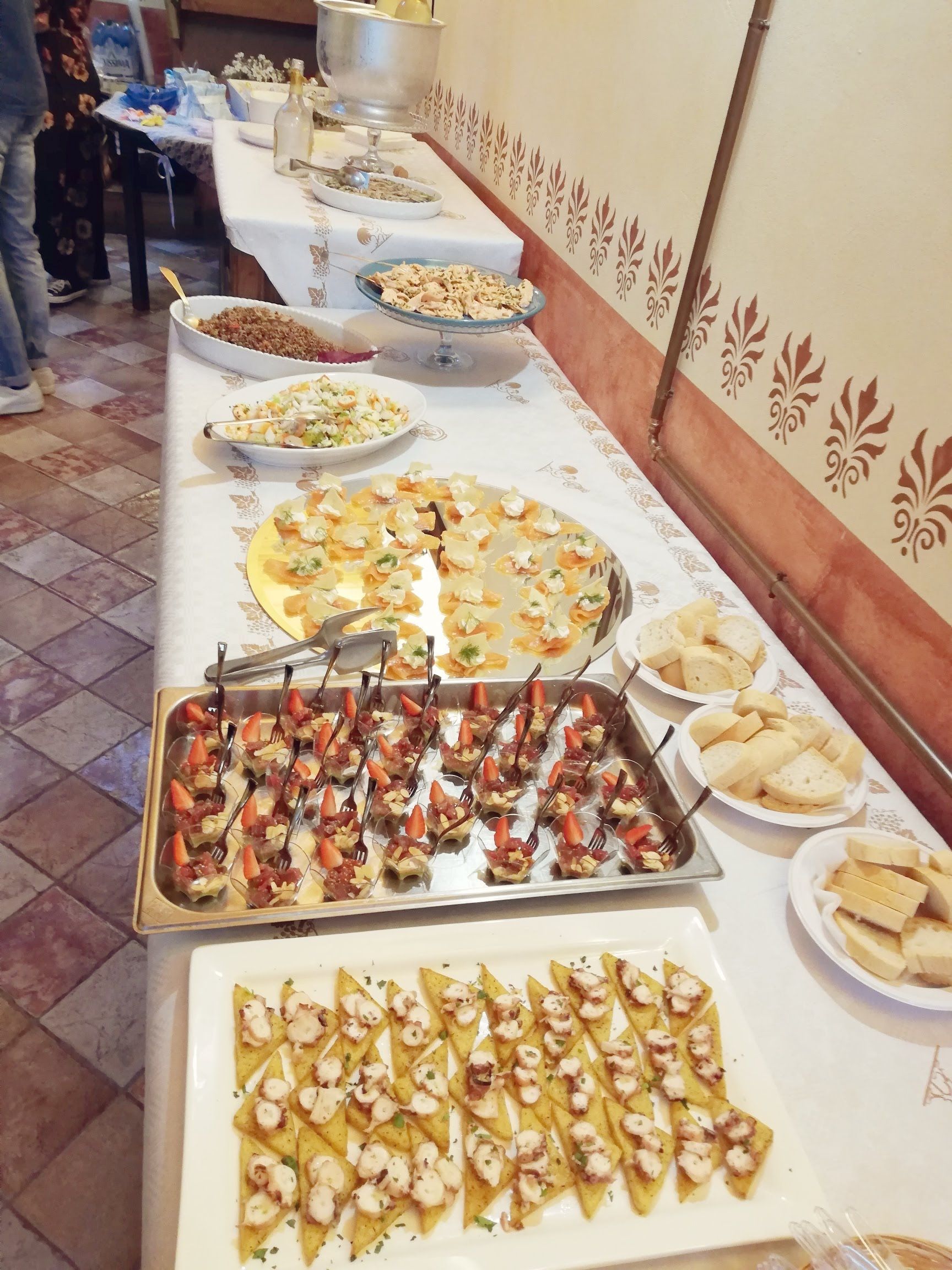 Tavolo da buffet con un assortimento di antipasti e dessert su una tovaglia bianca, contro una parete decorata.