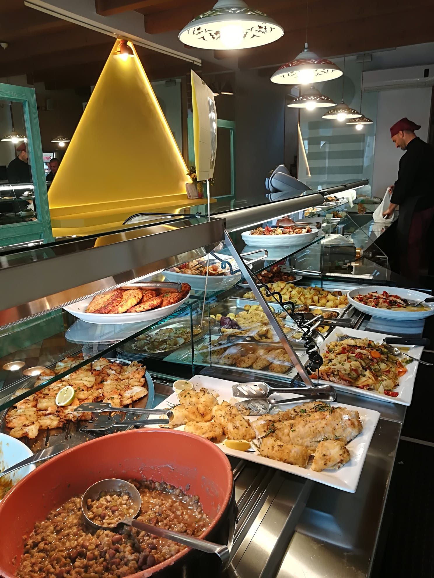 Tavolo da buffet con un assortimento di piatti caldi illuminati da luci soffuse, con una persona che serve il cibo sullo sfondo.