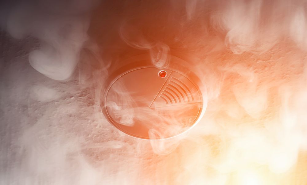 Close-up of a circular smoke detector on a ceiling, surrounded by wisps of smoke and orange light — Instant Spark Electrical in Glen Isla, QLD