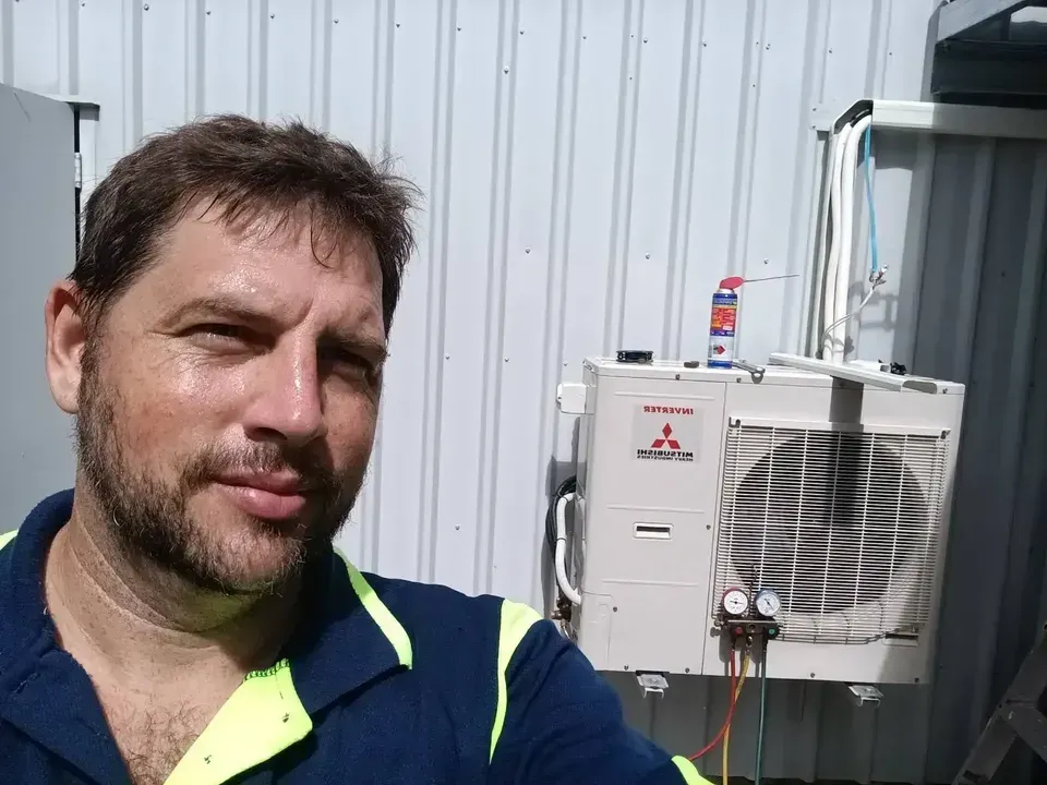 A Man Is Looking At The Camera With A Air Conditioning Unit Behind  — Instant Spark Electrical in Glen Isla, QLD