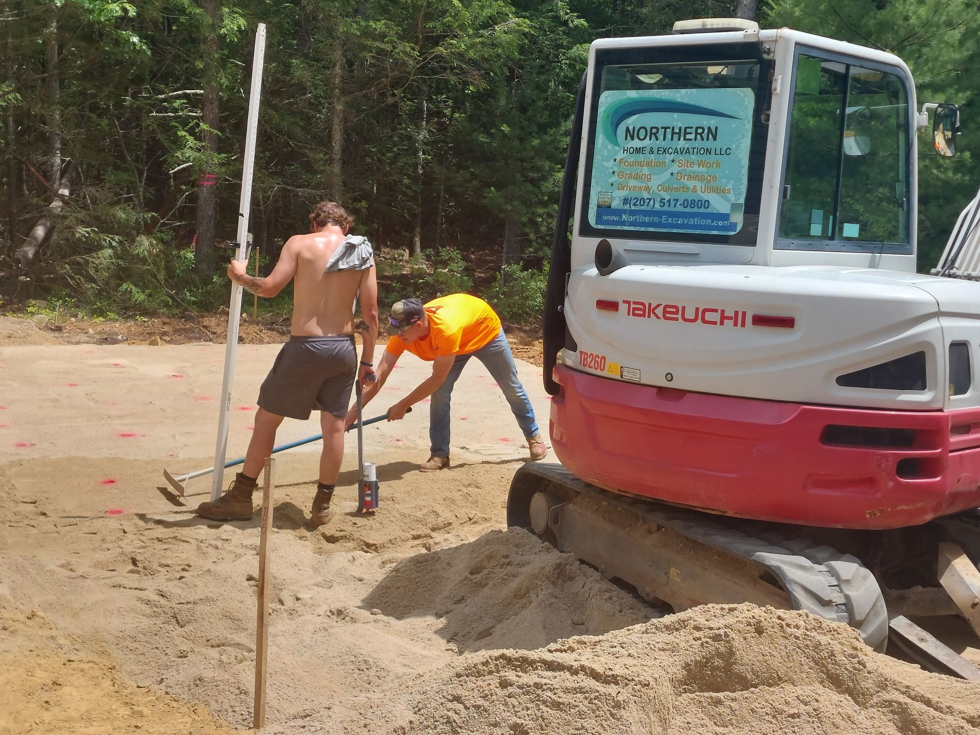 Two men are digging in the dirt in front of a takeuchi excavator.