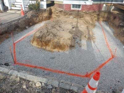 A construction site with a brick house in the background and a large hole in the ground.
