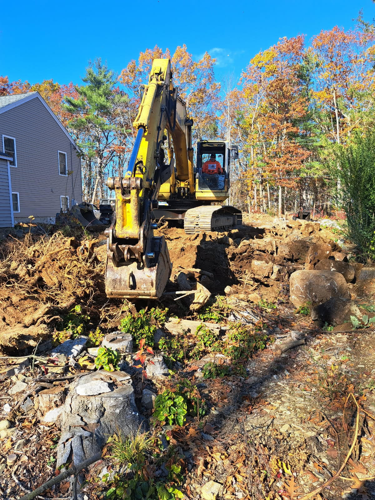 Yellow excavator digging into earth near a house; fall foliage in background.