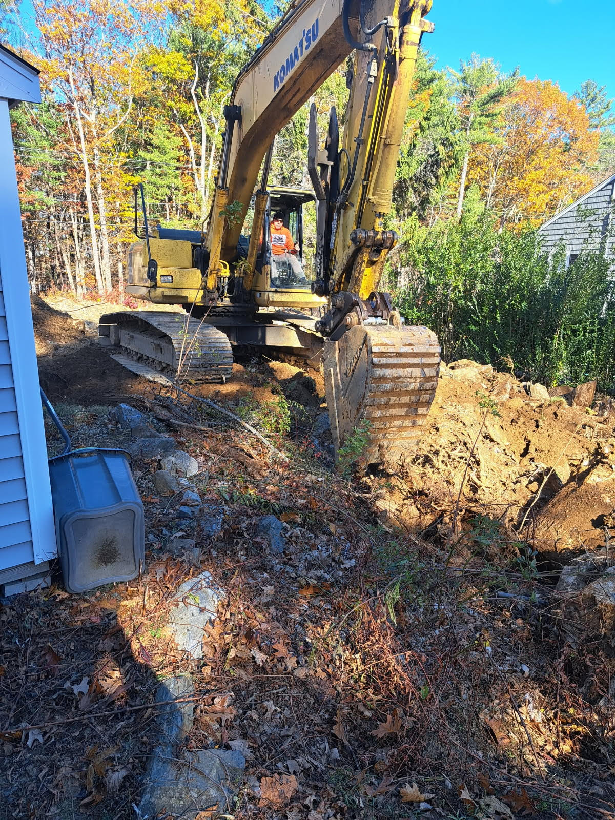 Yellow excavator digging along side of a light blue house. Trees and foliage surround the construction site.