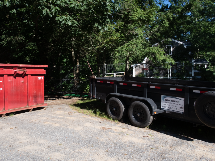A dumpster and a trailer are parked in a gravel lot.